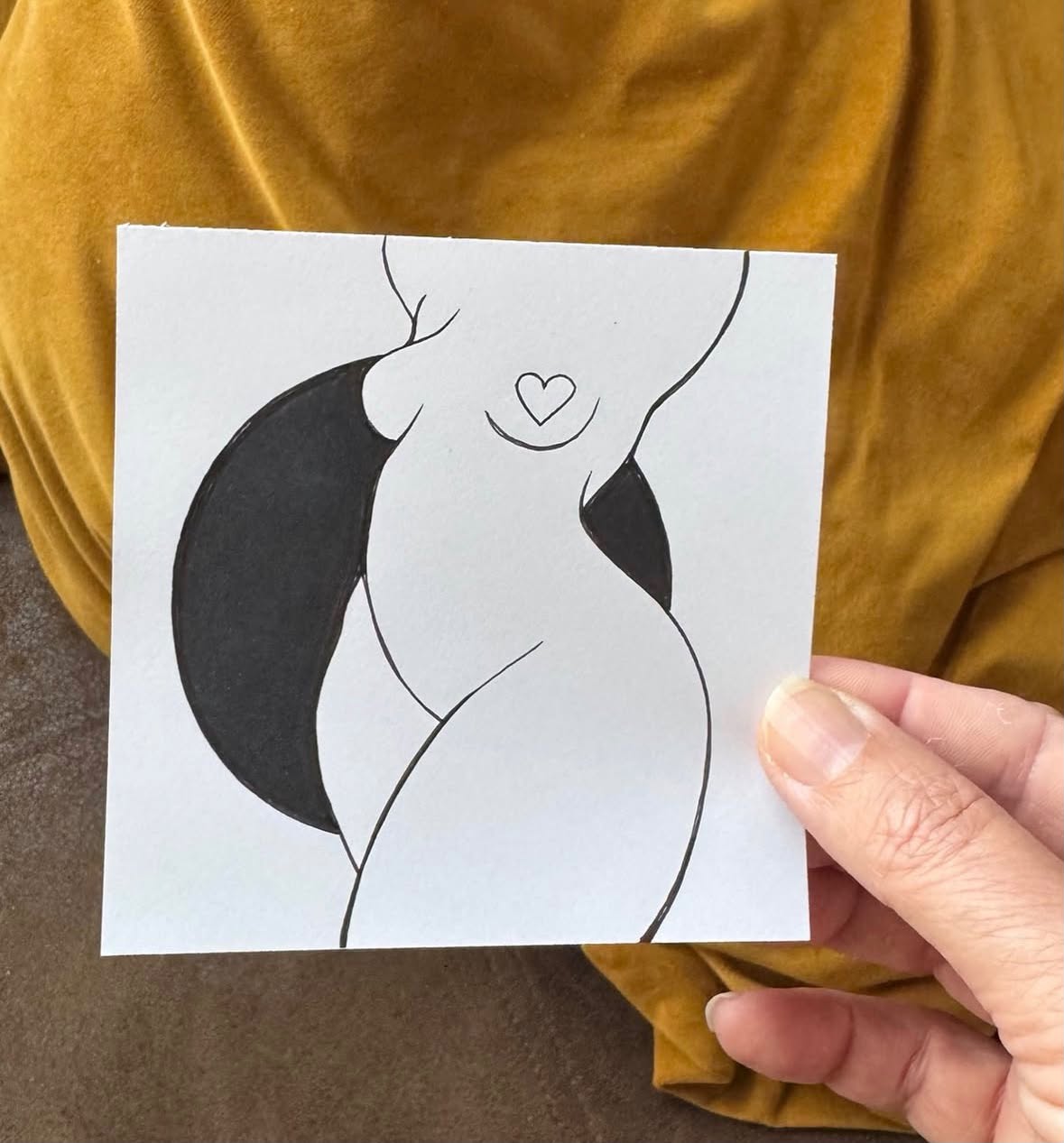 Hand houdt een zwart-witte Being-tekening van Hanneke Wetzer vast, voor een okergele achtergrond.